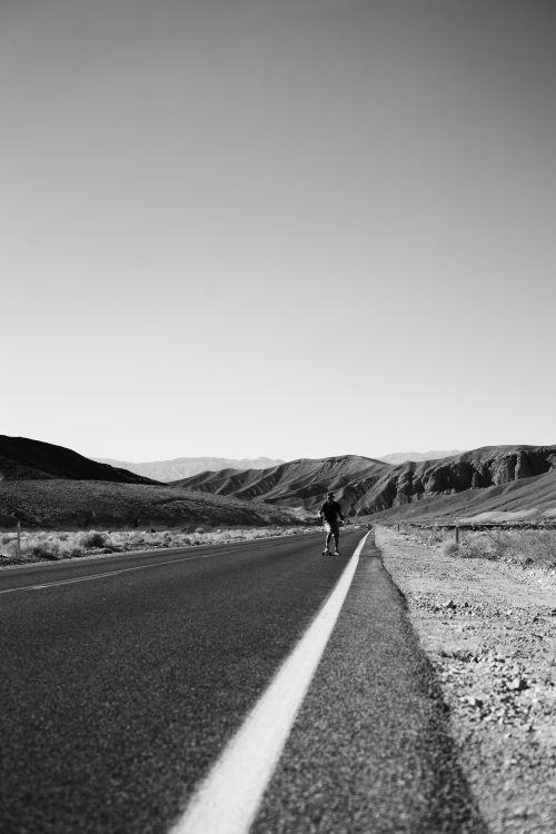 Free Black and white image of a lone man walking on a deserted road through mountainous terrain. Stock Photo