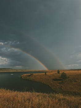 A breathtaking view of a double rainbow stretching over a tranquil field and river, post-rain.