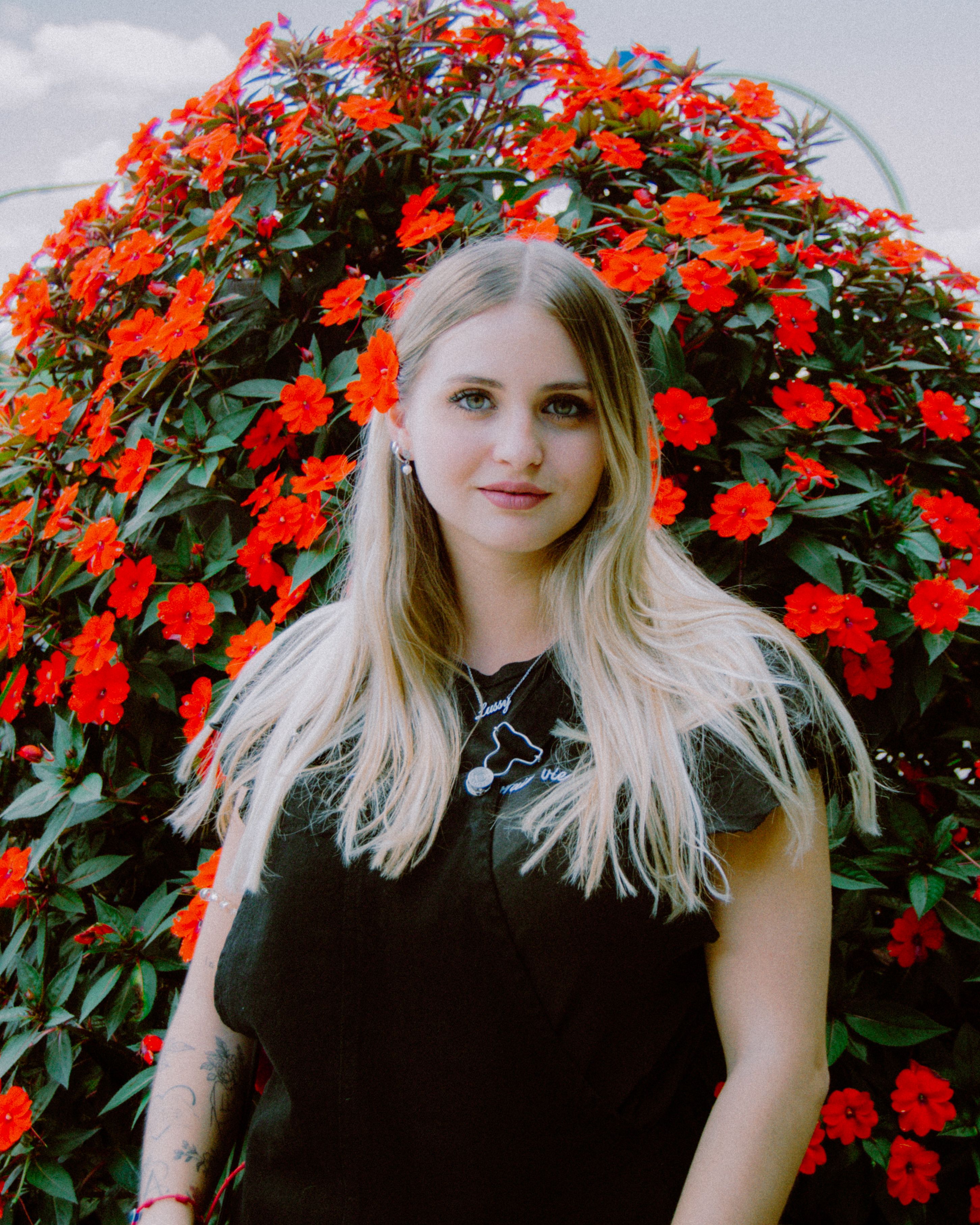 Blonde woman in black dress poses in front of vibrant red flowers, outdoors.