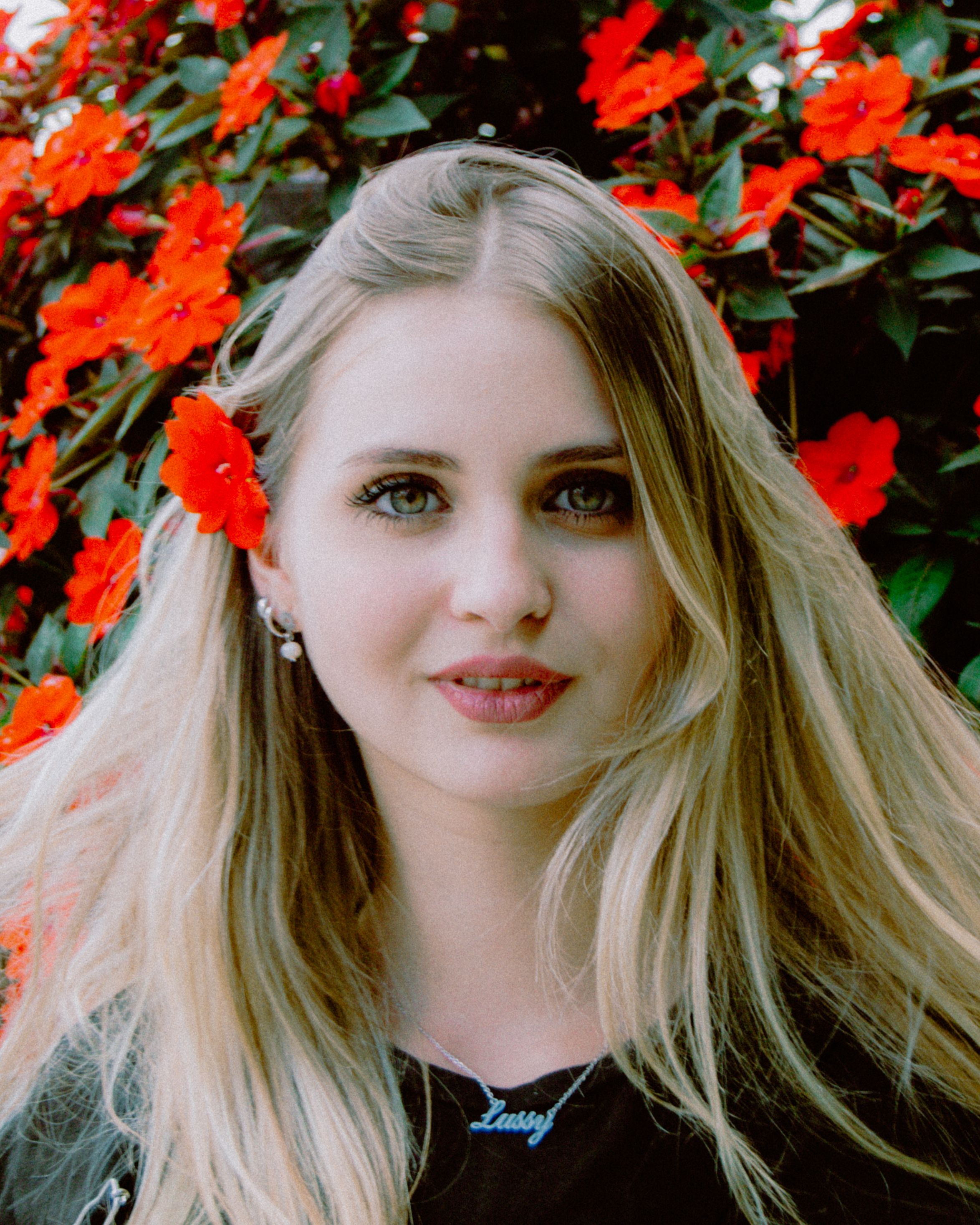 Young blonde woman smiling outdoors with red flowers in her hair, creating a vibrant summer portrait.