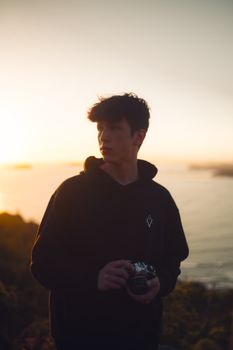 A young male photographer holding a camera, standing by a beach at sunset.