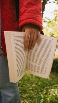 A person in a red jacket holds an open book outdoors on a sunny day. Focus on the hand and book.