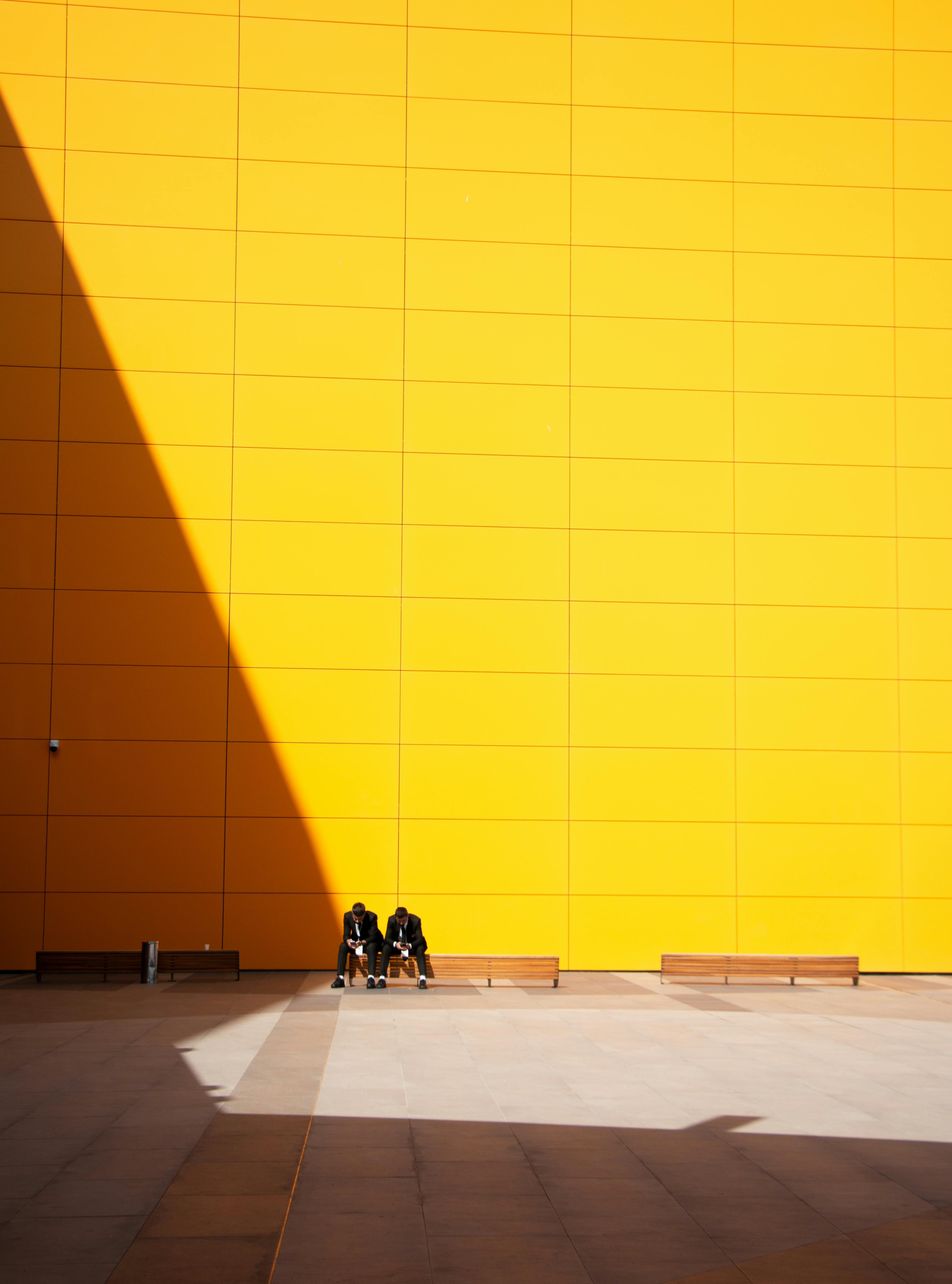 Two people sitting by a striking yellow wall in Pristina, creating a minimalist urban scene.