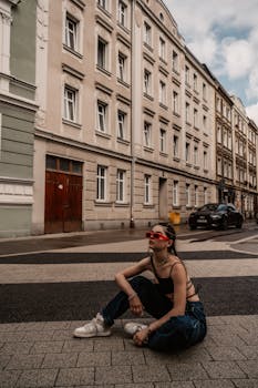Fashionable young woman with sunglasses sits on city street, surrounded by vintage architecture.