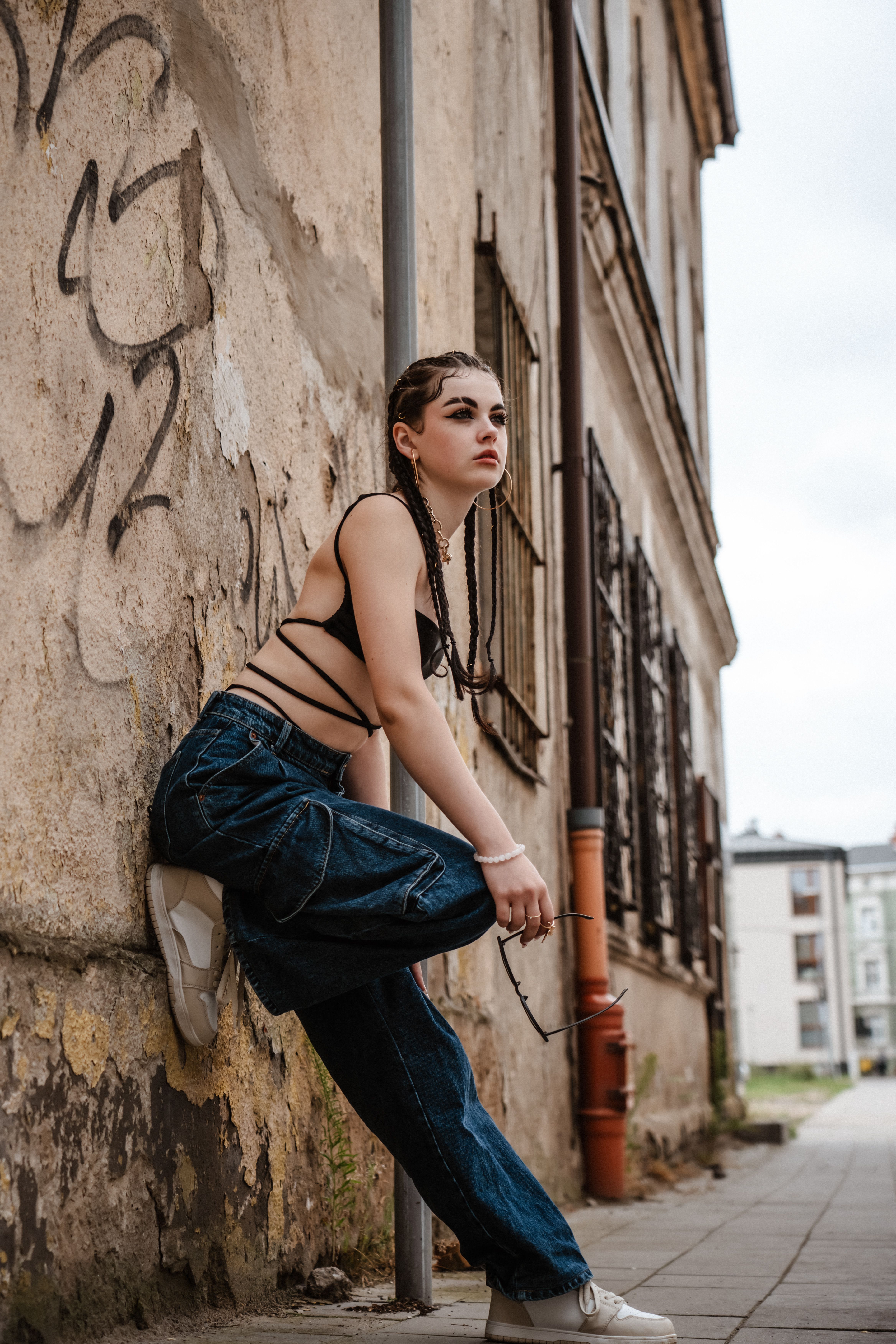 Stylish young woman in streetwear leans on graffiti wall, urban setting.