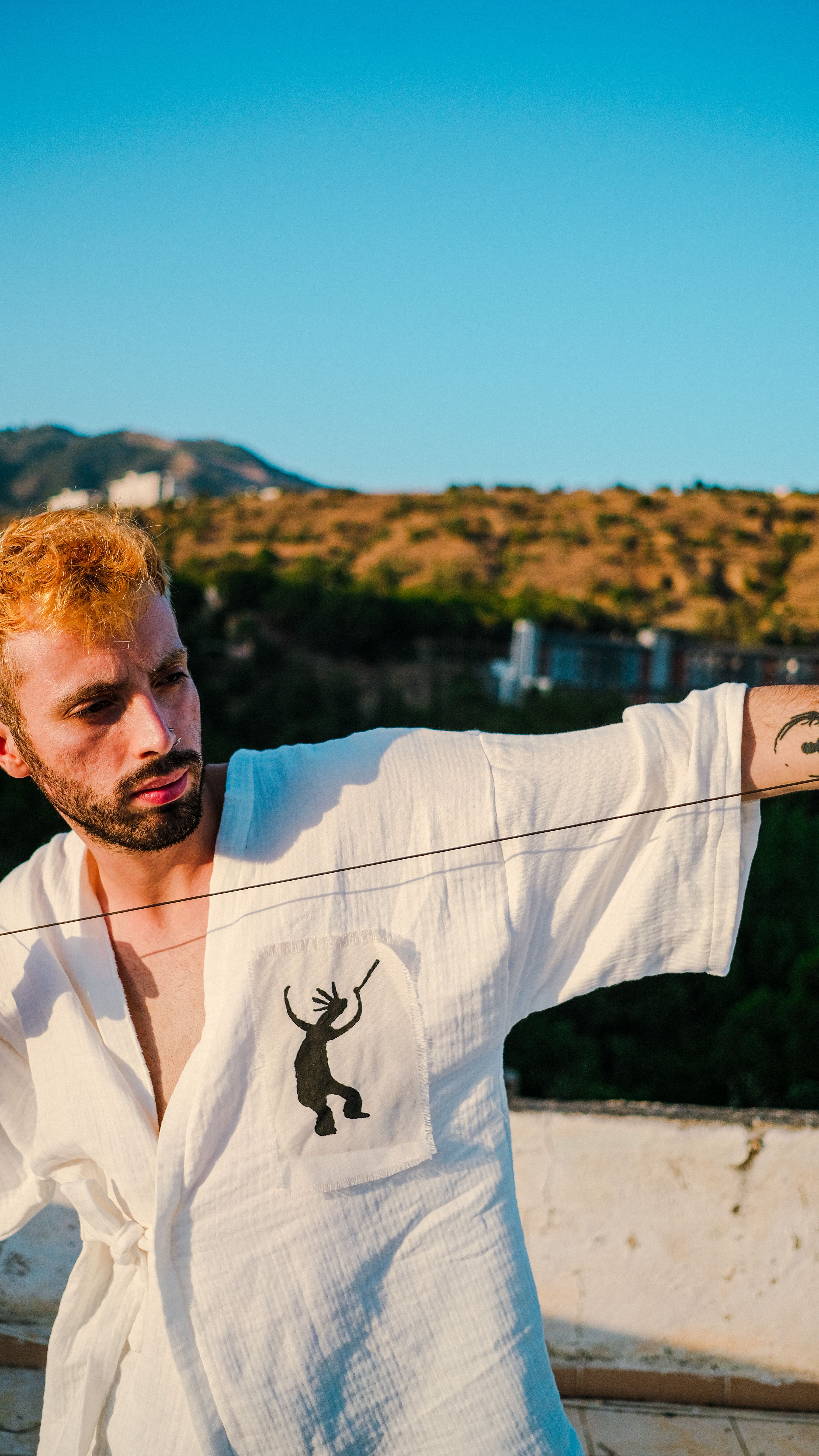 A young man in a white robe poses outdoors with mountains in the background.
