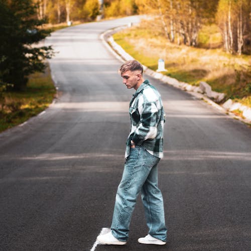 Young Man in Jeans and Checkered Shirt Standing on an Asphalt Road 
