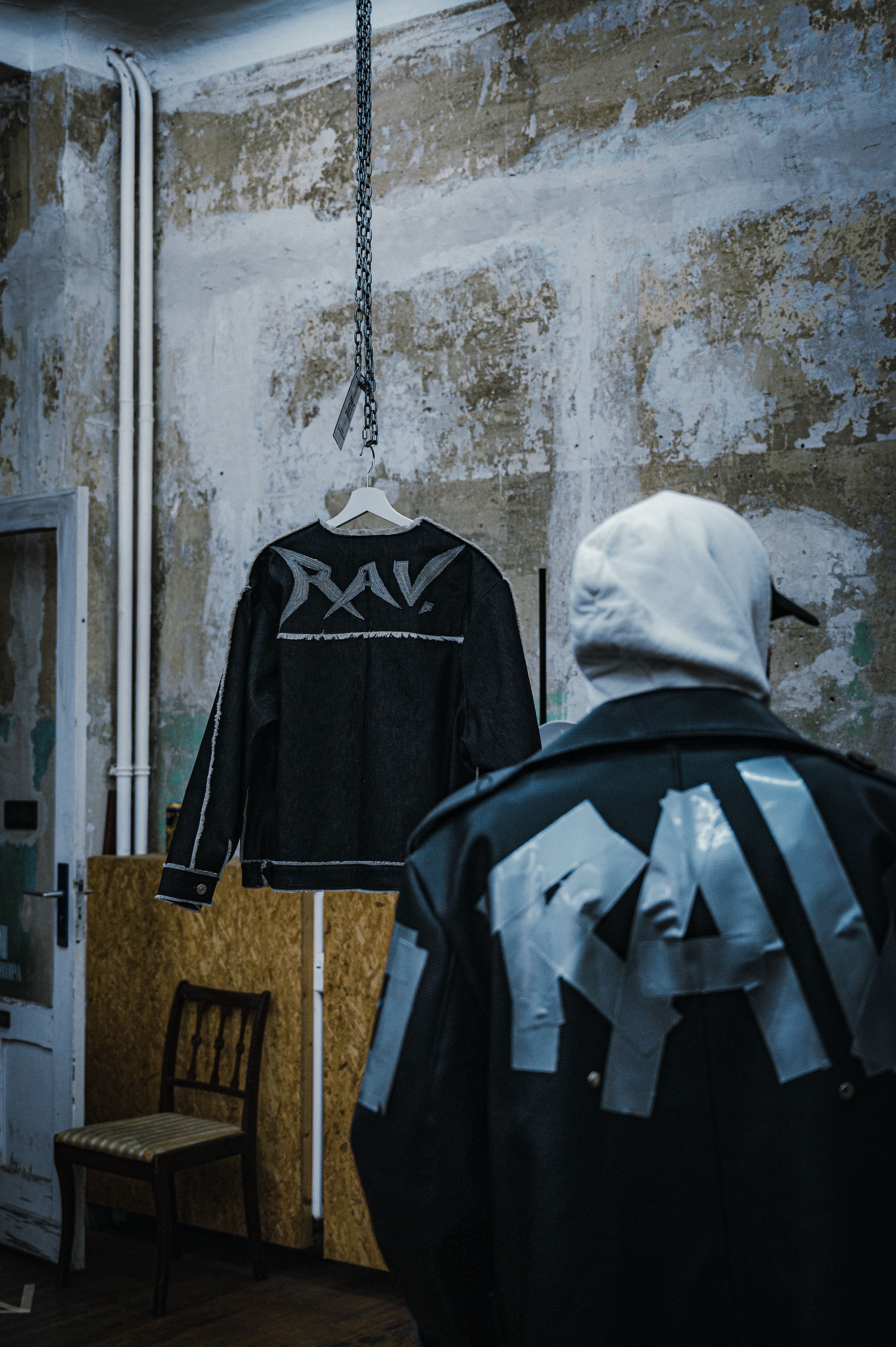 A fashion model in a hoodie and leather jacket examines a hanging garment in an urban industrial setting.