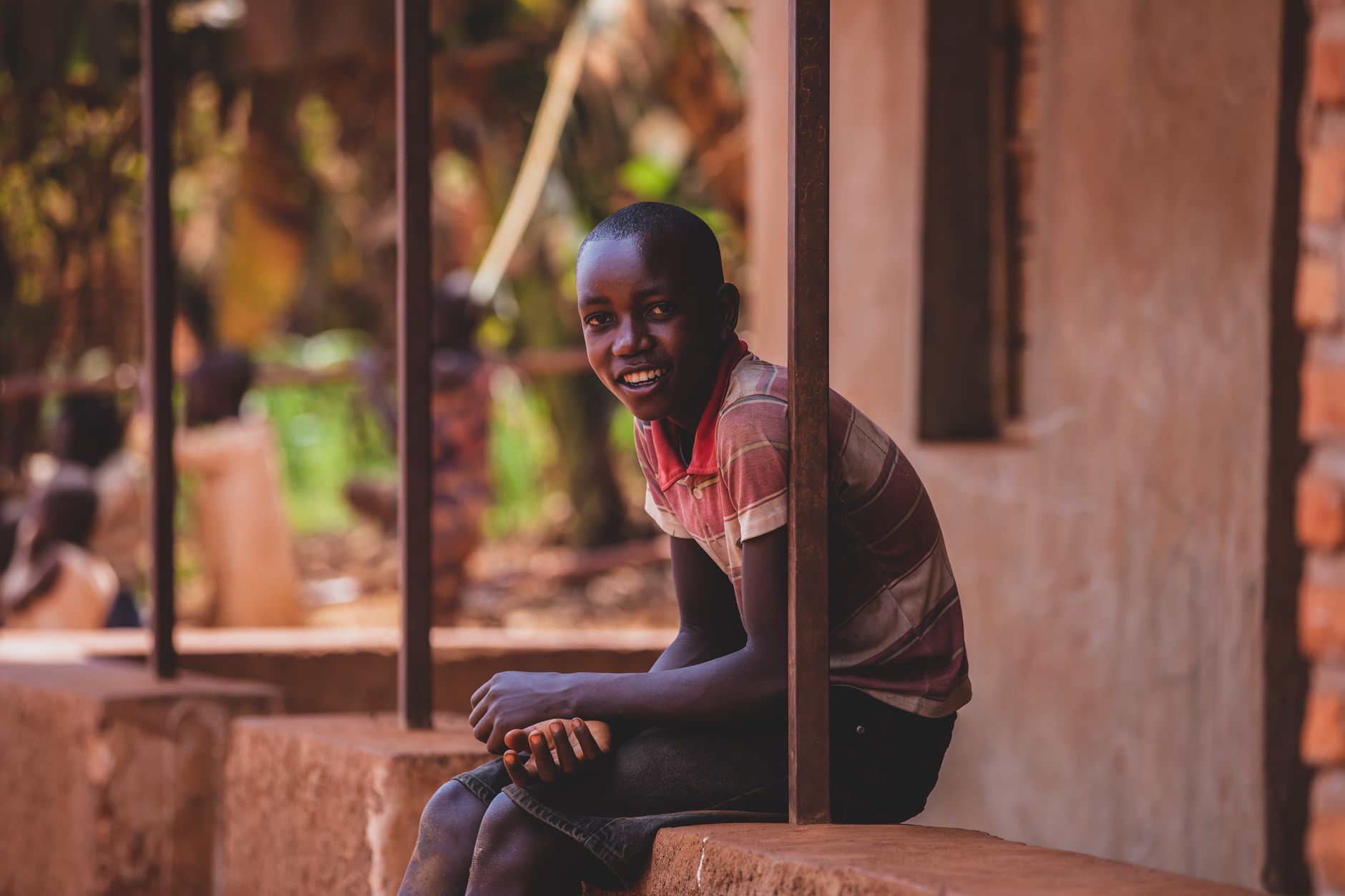 A young boy sits outdoors, smiling warmly on a rural porch, against a blurred background.