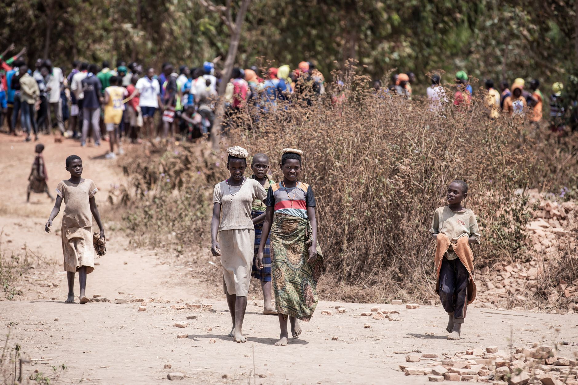 Women and children walk along a dirt road in a rural African village, showcasing everyday life.