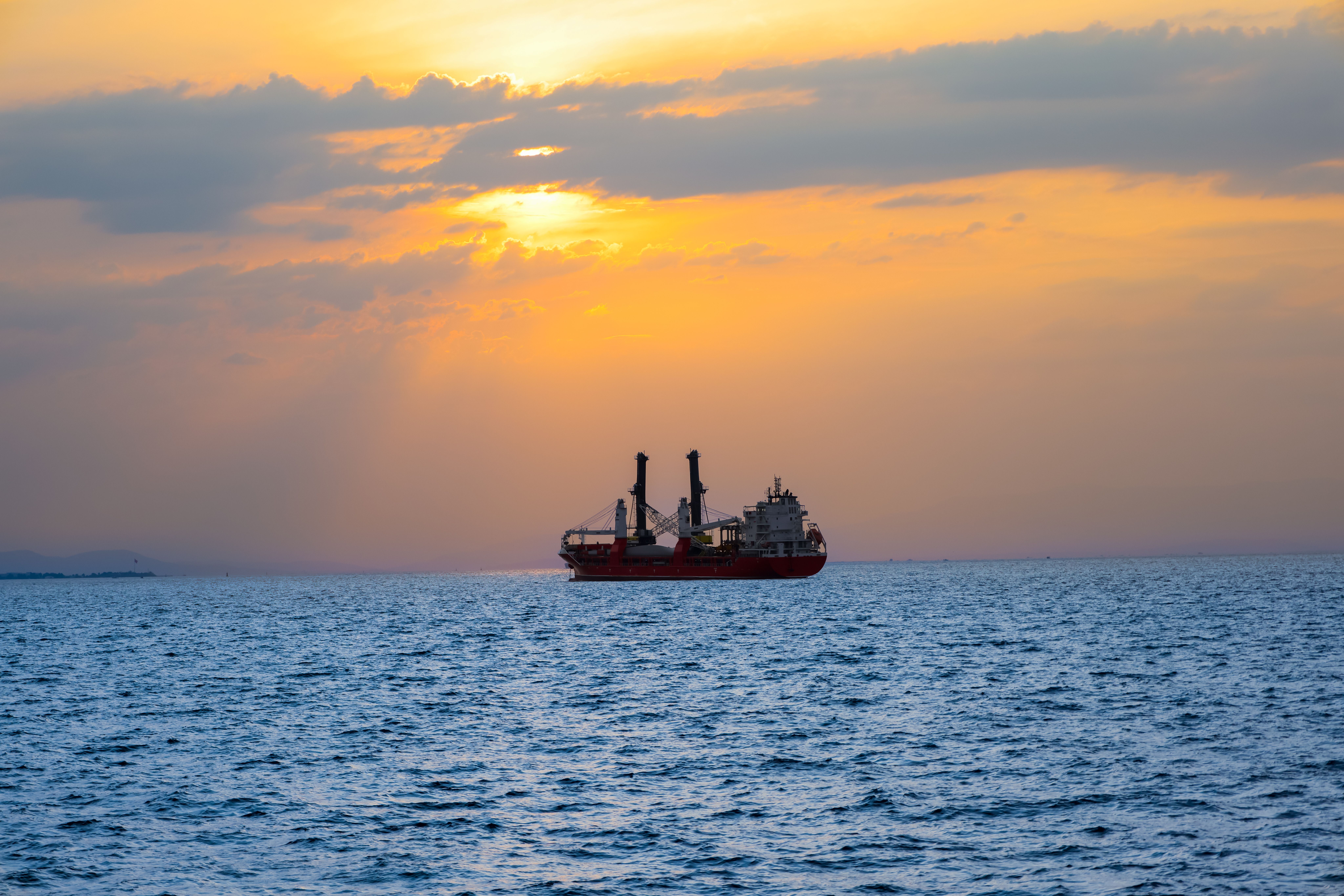 A cargo ship sails across the ocean against a vibrant sunset, creating a serene silhouette.