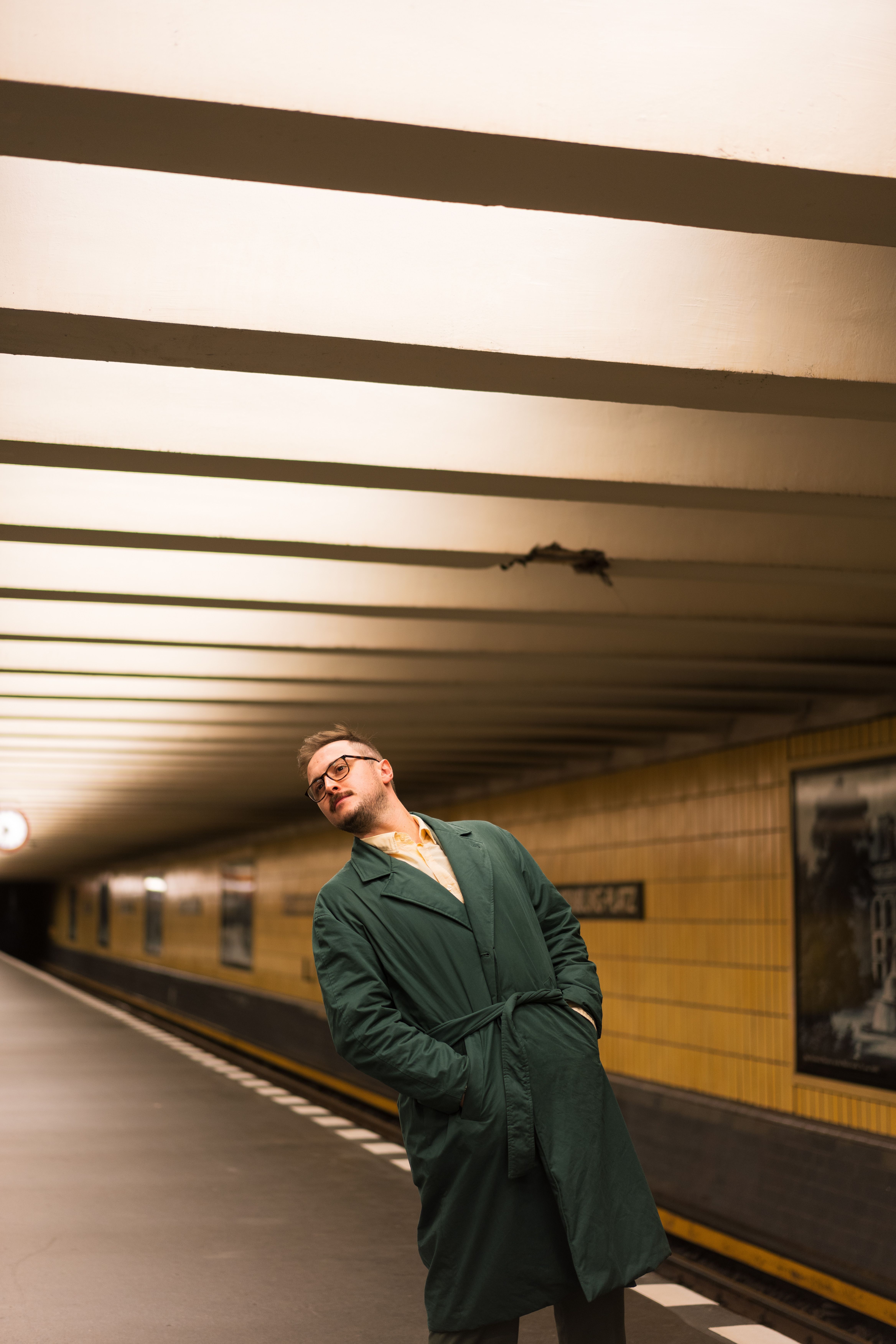 Man in a green coat posing in a Berlin subway station, urban fashion scene.