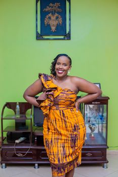 Smiling African woman in a bold orange dress posing indoors, showcasing vibrant fashion.