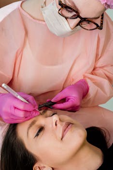 Beautician in pink attire provides eyebrow treatment to a relaxed client in a professional setting.