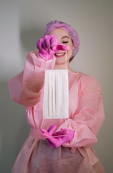 A female healthcare professional holding a surgical mask, smiling while wearing pink protective gear.