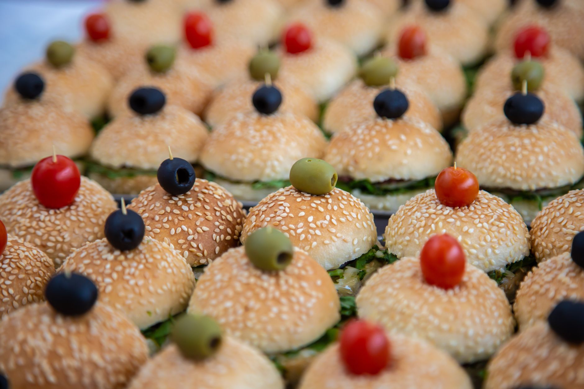 Close-up of mini burgers topped with olives and cherry tomatoes on sesame buns.