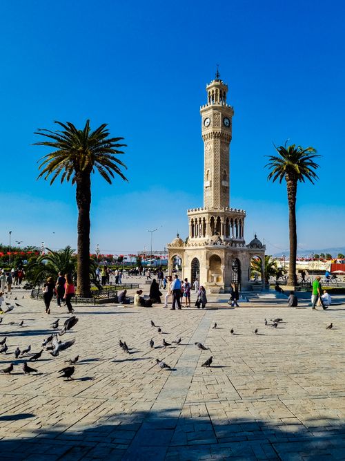 Free The iconic İzmir Clock Tower stands tall amid bustling crowds and palm trees on a sunny day. Stock Photo