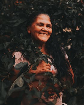 一位留着长长黑发的老年妇女，在户外茂密的绿植环绕中快乐地微笑着