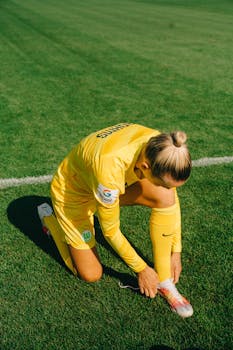A female soccer player in yellow sportswear tying her shoelaces on a grass field.