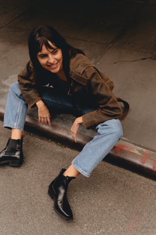 Woman in Brown Leather Jacket, Blue Jeans, and Black Boots Sitting on a Curb