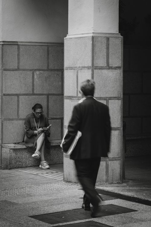 Free A monochrome cityscape featuring a woman reading and a man walking, captures daily urban life. Stock Photo