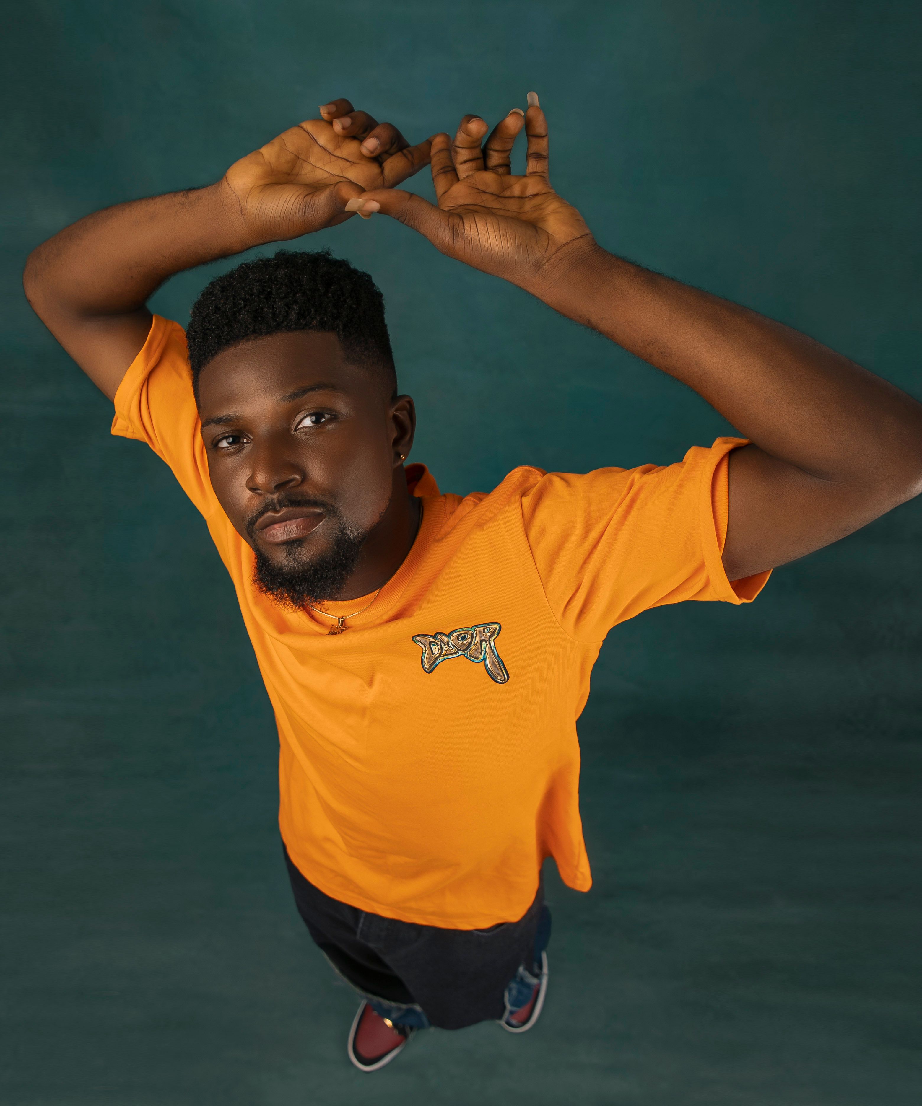 Fashionable portrait of a man posing in an orange t-shirt, arms raised, studio setting.