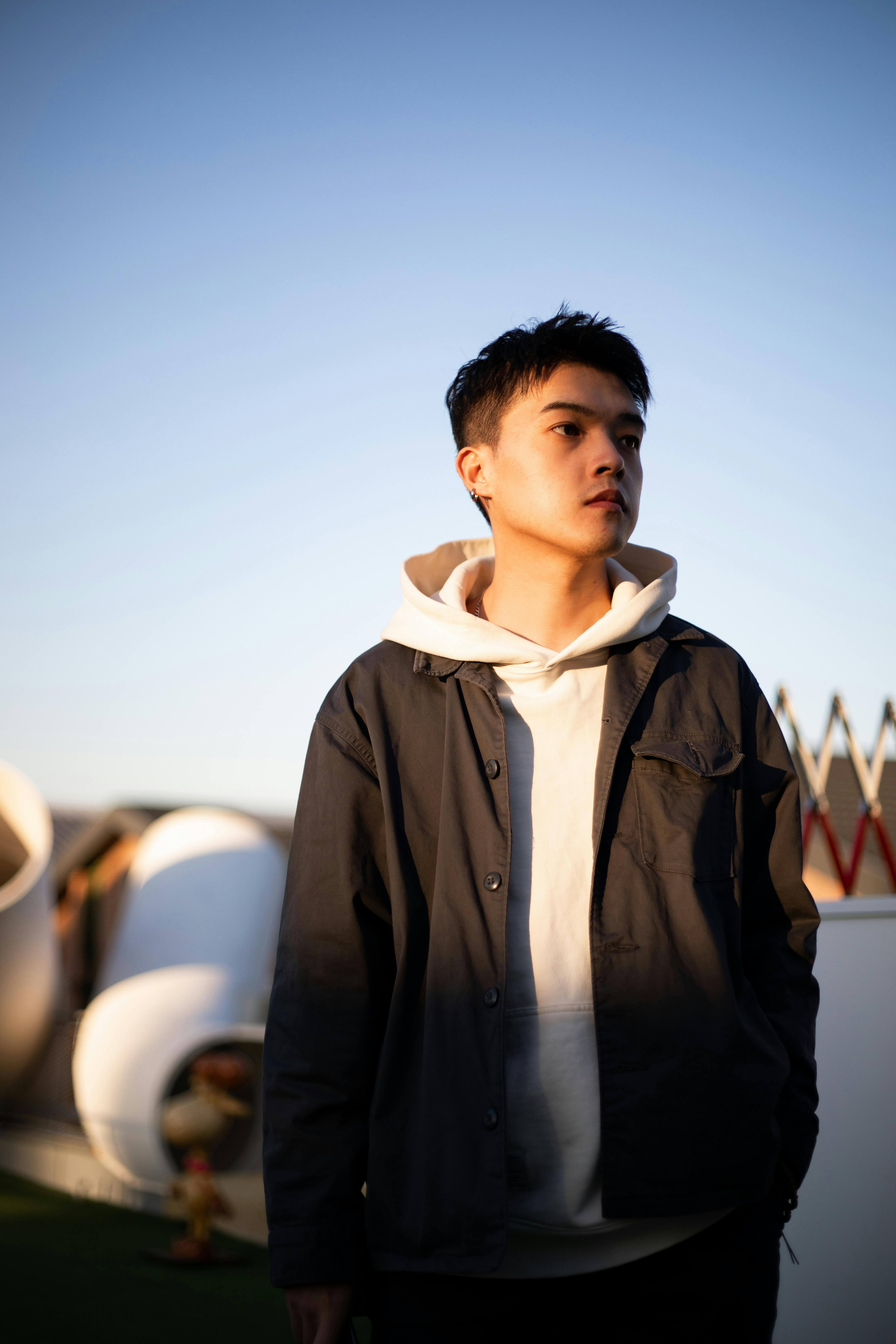 A young man in a black shacket and white hoodie poses outdoors in a stylish urban environment.