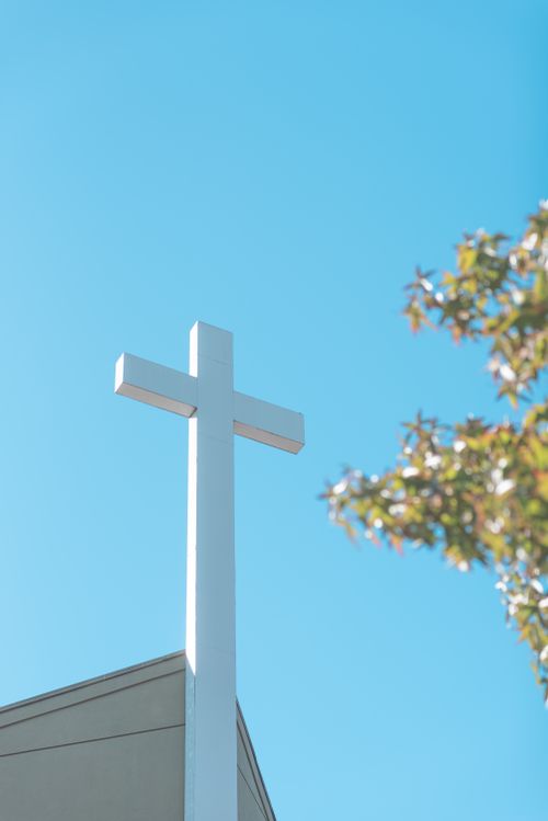 Free A white church cross stands against a bright blue sky with tree branches in view. Stock Photo
