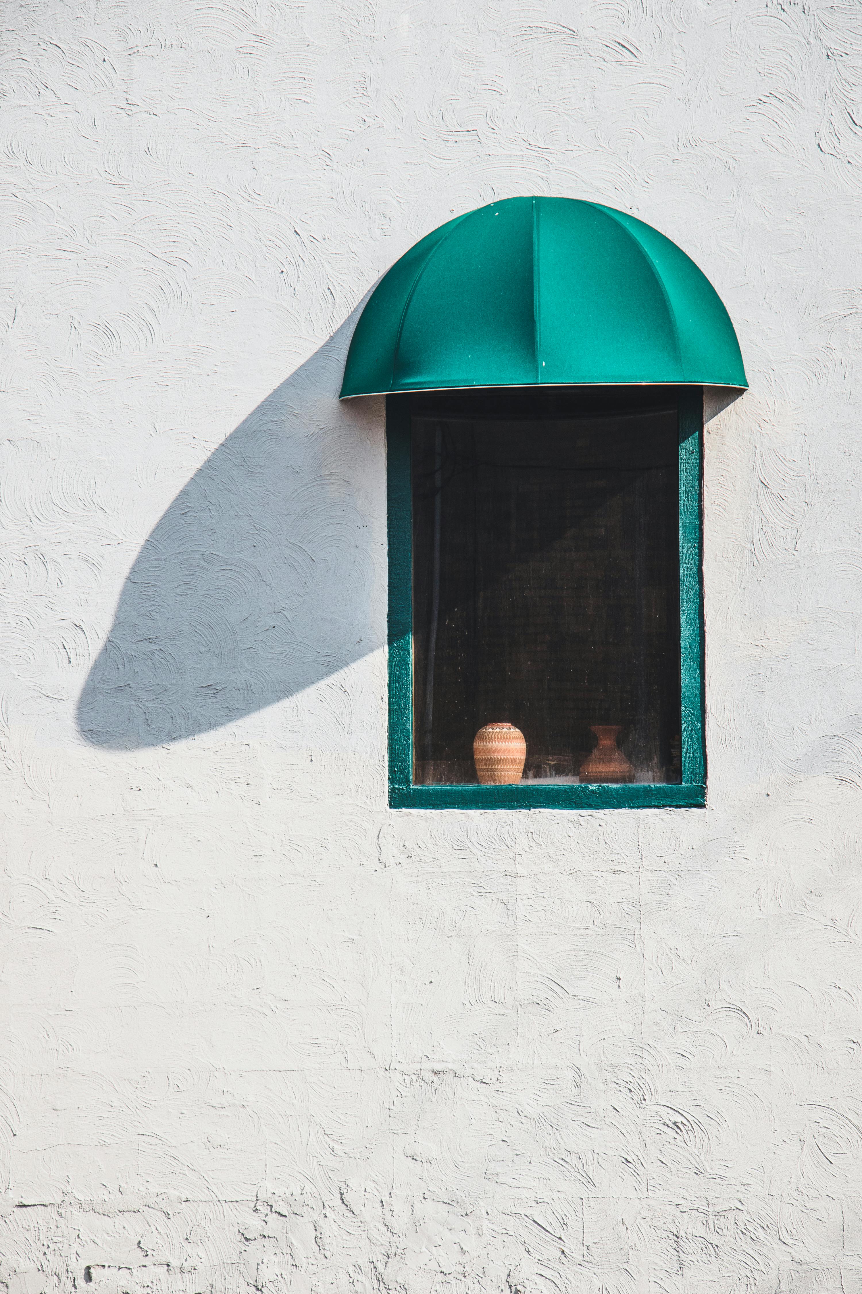 Aesthetic window with clay pots and green awning casting a shadow on a white wall.