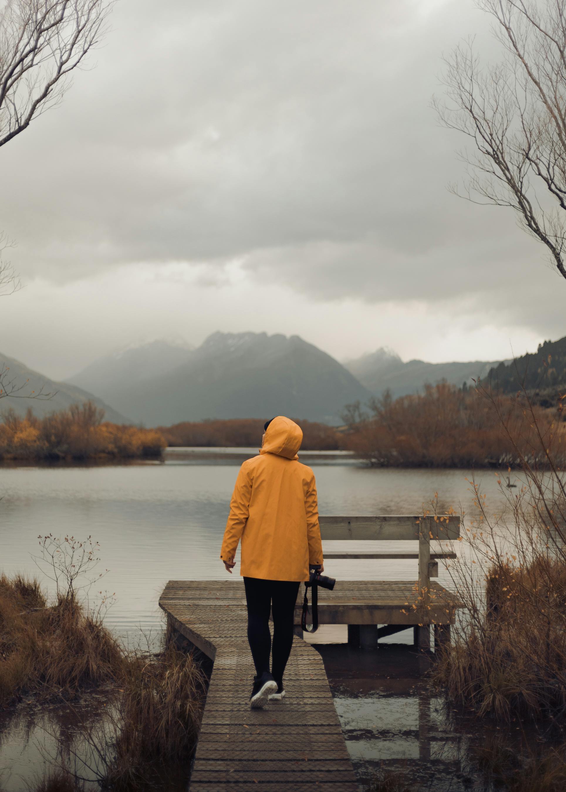 A person in a yellow raincoat walks on a jetty at Lake Wakatipu, surrounded by autumn scenery in Queenstown, New Zealand.