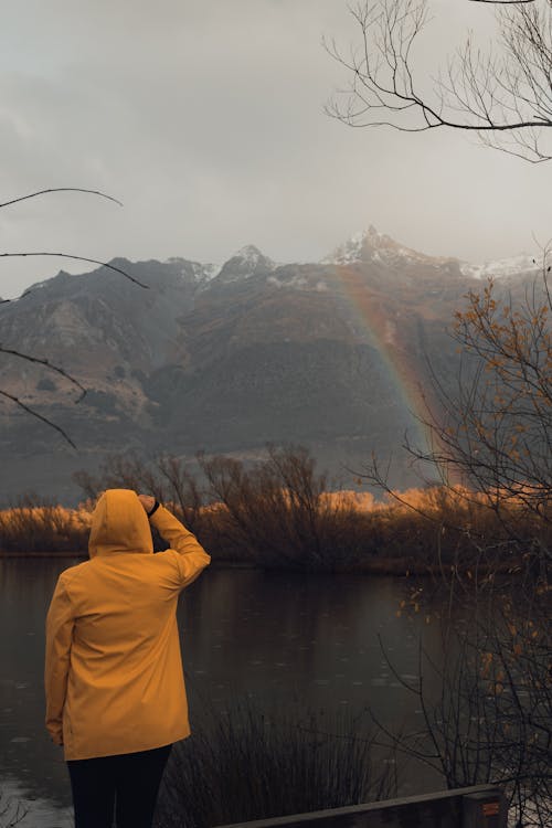 Tourist in a Yellow Raincoat Standing in the Rain Watching a Rainbow over the Lake