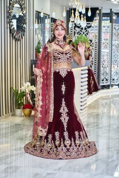 A beautiful bride wearing an ornate burgundy and gold traditional caftan indoors.
