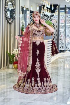 A glamorous bride in a richly adorned maroon wedding dress with gold details, indoor setting.