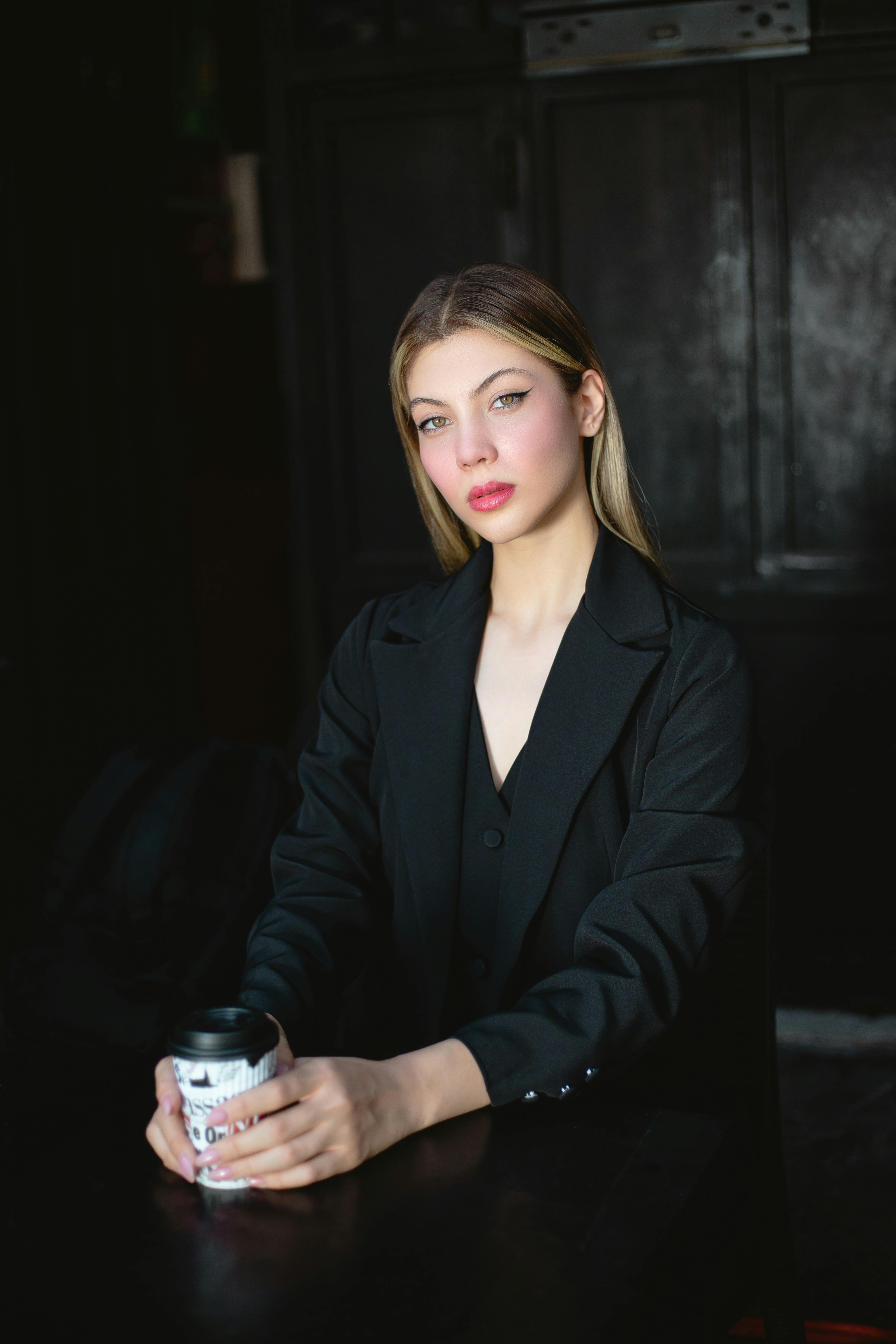 Elegant young woman in black coat holding a coffee cup while seated indoors.