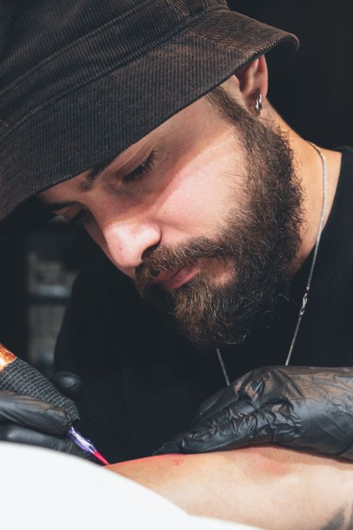 Free Focused tattoo artist with beard and bucket hat creating body art in a studio. Stock Photo