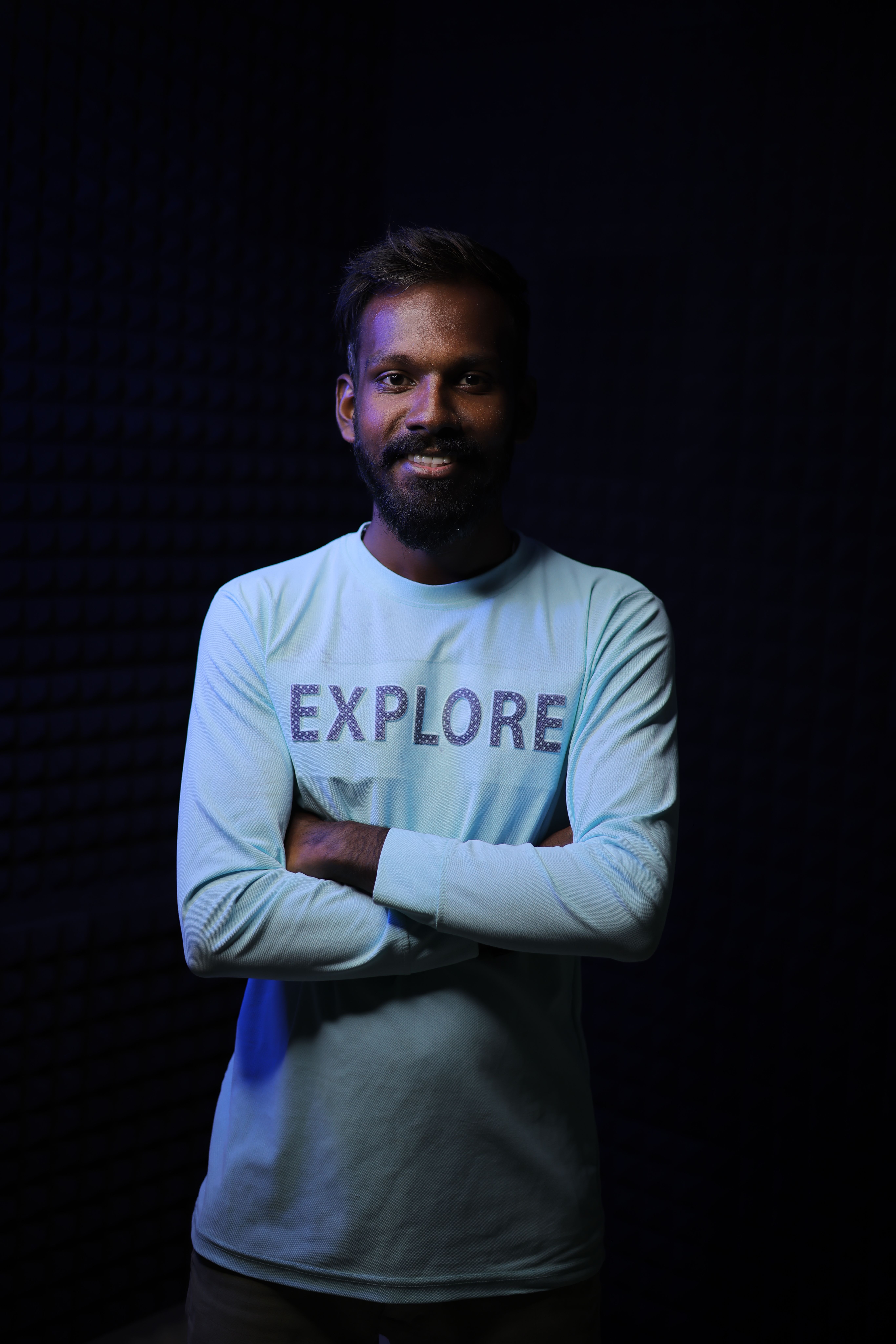 A smiling man stands confidently in a black studio background wearing an 'Explore' sweatshirt.