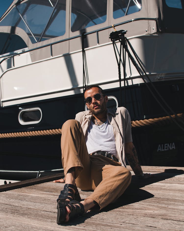 Man In Sandy Chino Pants, Beige Shirt, And Sunglasses Posing On A Pier 