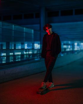 Young adult skateboarder standing in an urban setting under dramatic colorful lighting at night.