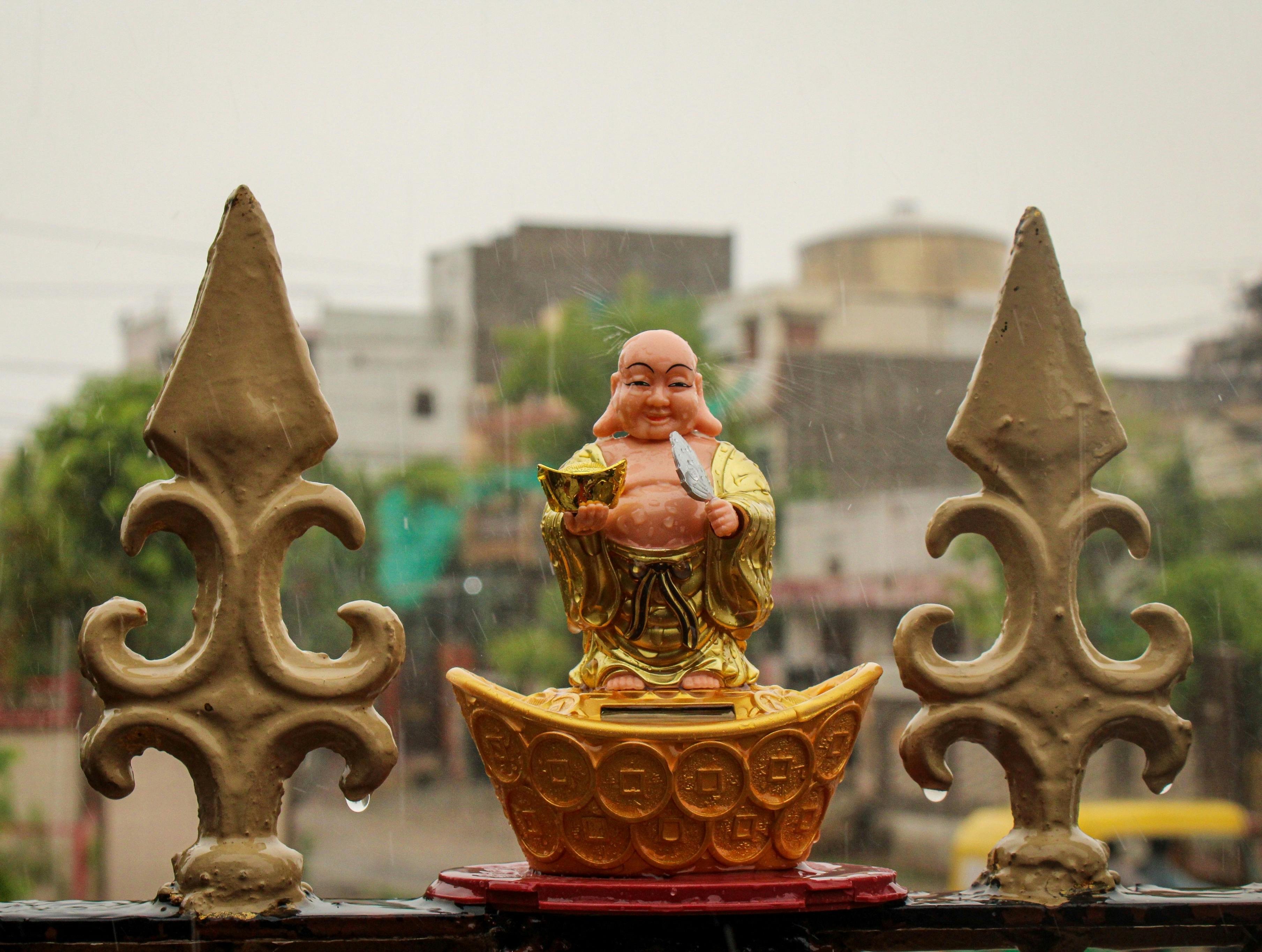 Smiling golden Buddha figurine on a rainy day with urban backdrop.