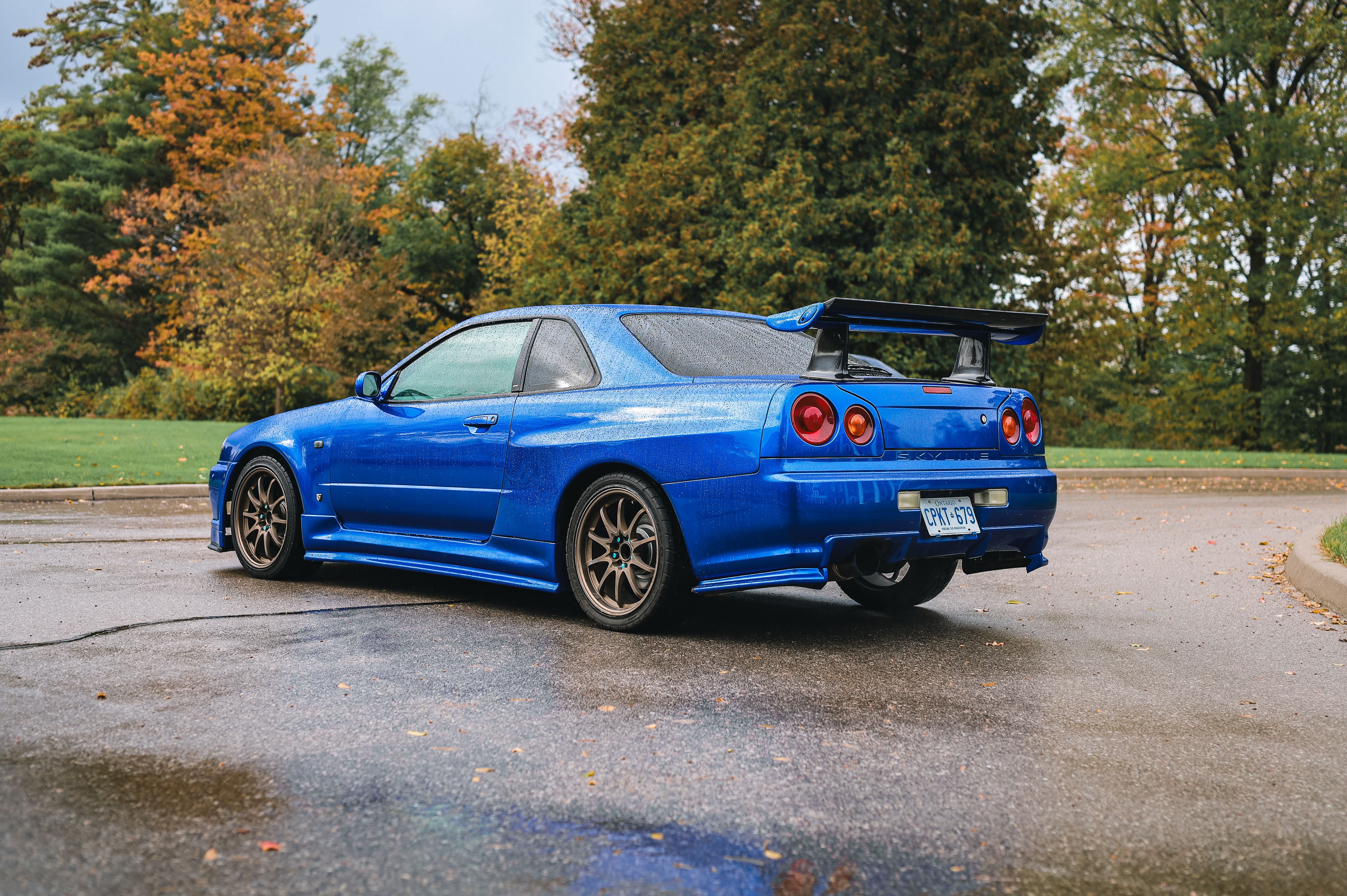 A vibrant blue Nissan Skyline GT-R parked on a wet road surrounded by autumn foliage.