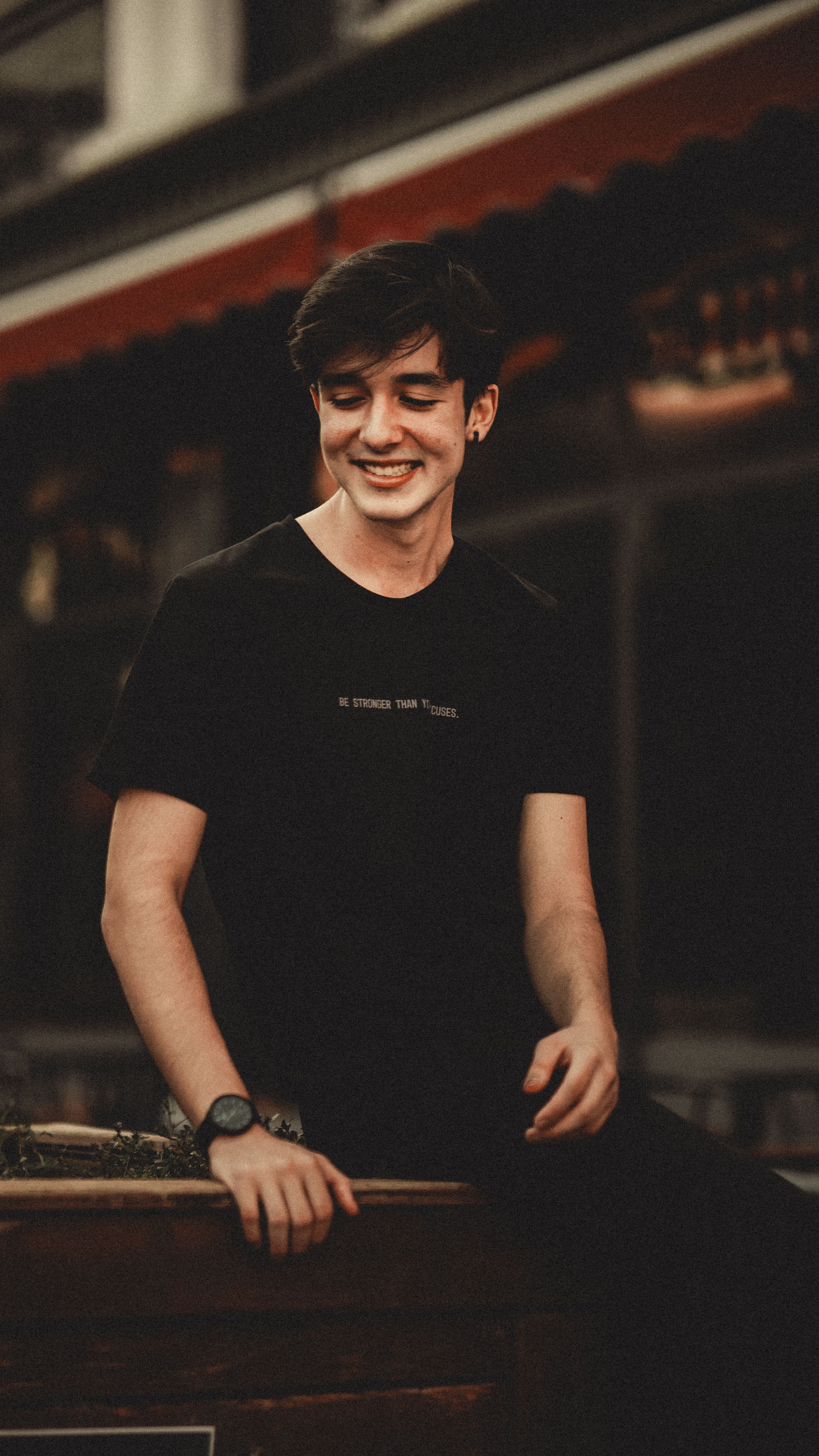 A joyful young man in a black shirt smiling outdoors, showcasing casual style in a city setting.