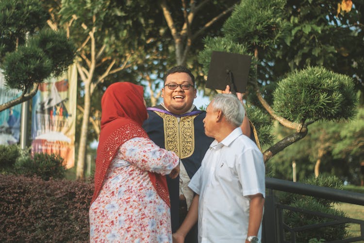 Man In A Gown And A Mortarboard Standing With His Parents And Smiling 