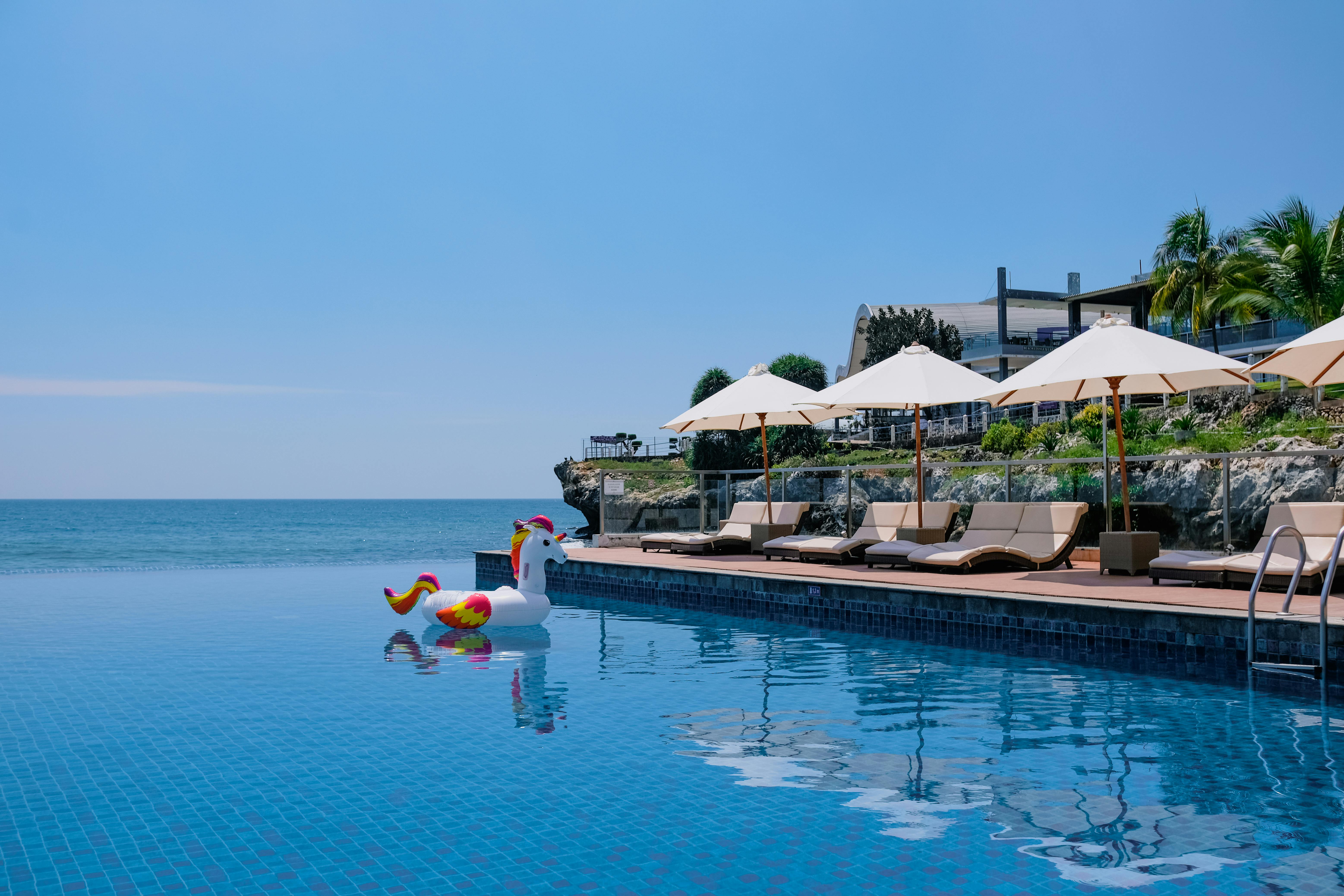 A tranquil beachside infinity pool with sunbeds, umbrellas, and a playful unicorn float under the summer sun.