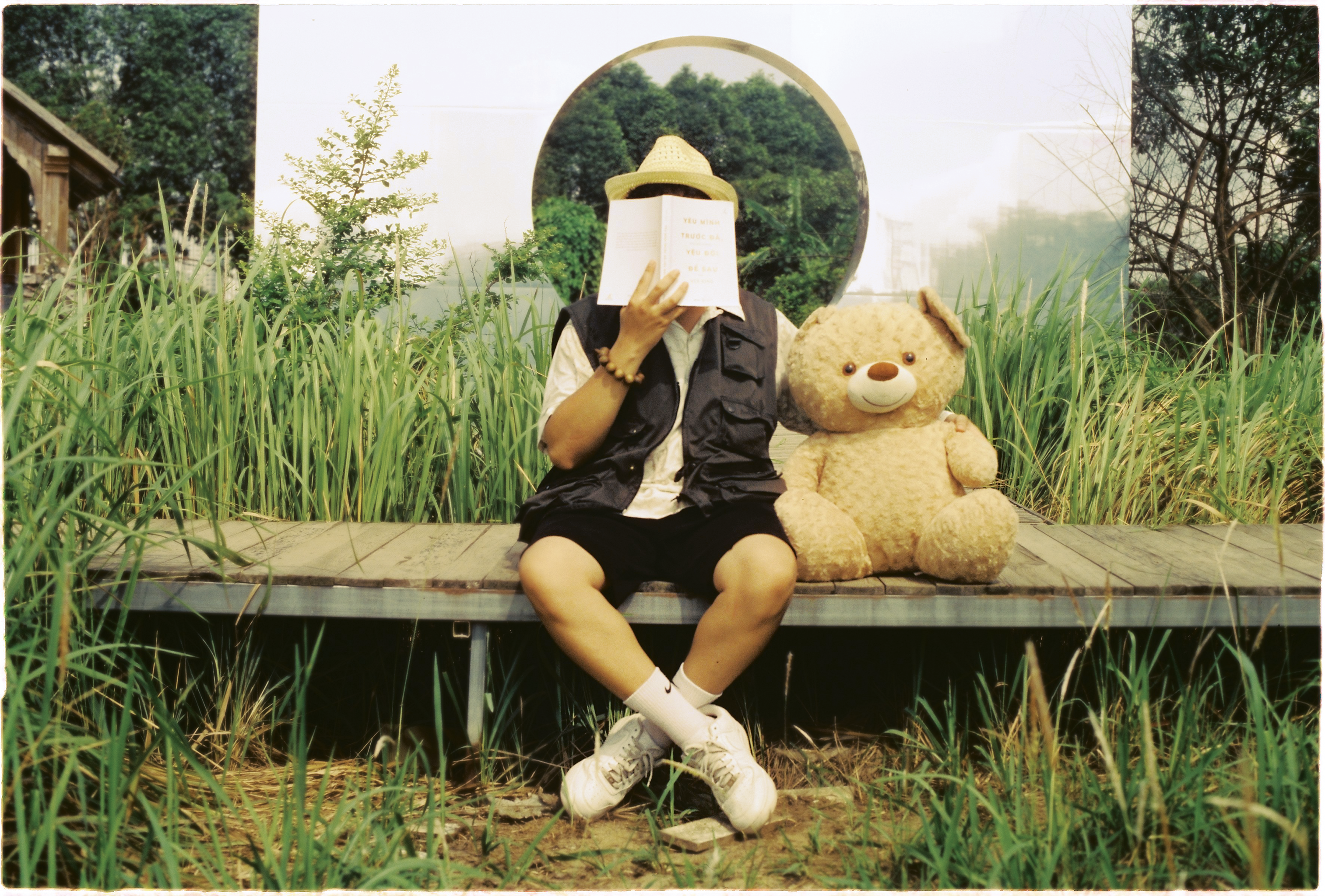 A young boy in a rural setting reads a book while sitting next to a plush teddy bear.