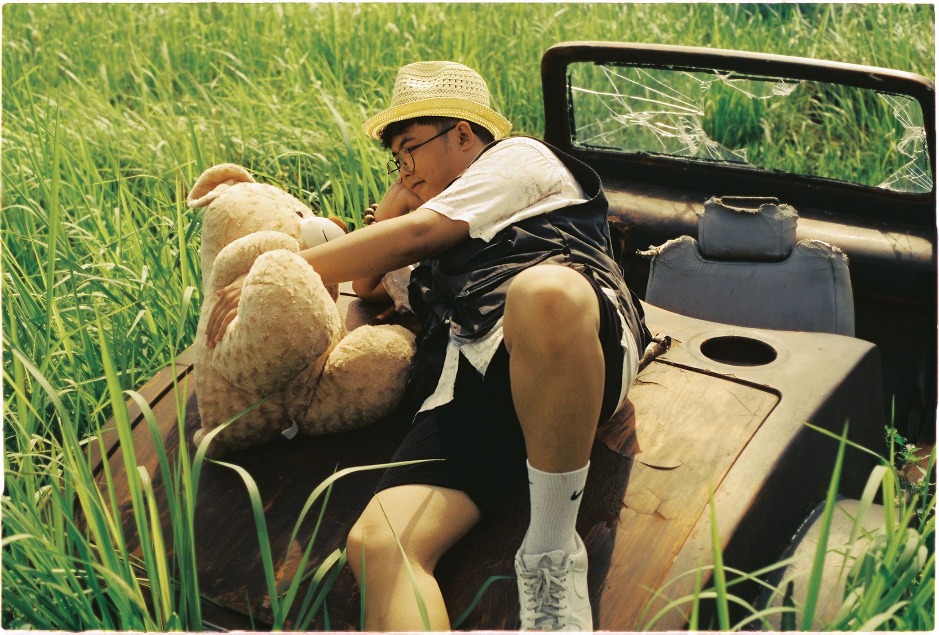 A man in a hat and glasses lying with a teddy bear in an overgrown, abandoned vehicle.