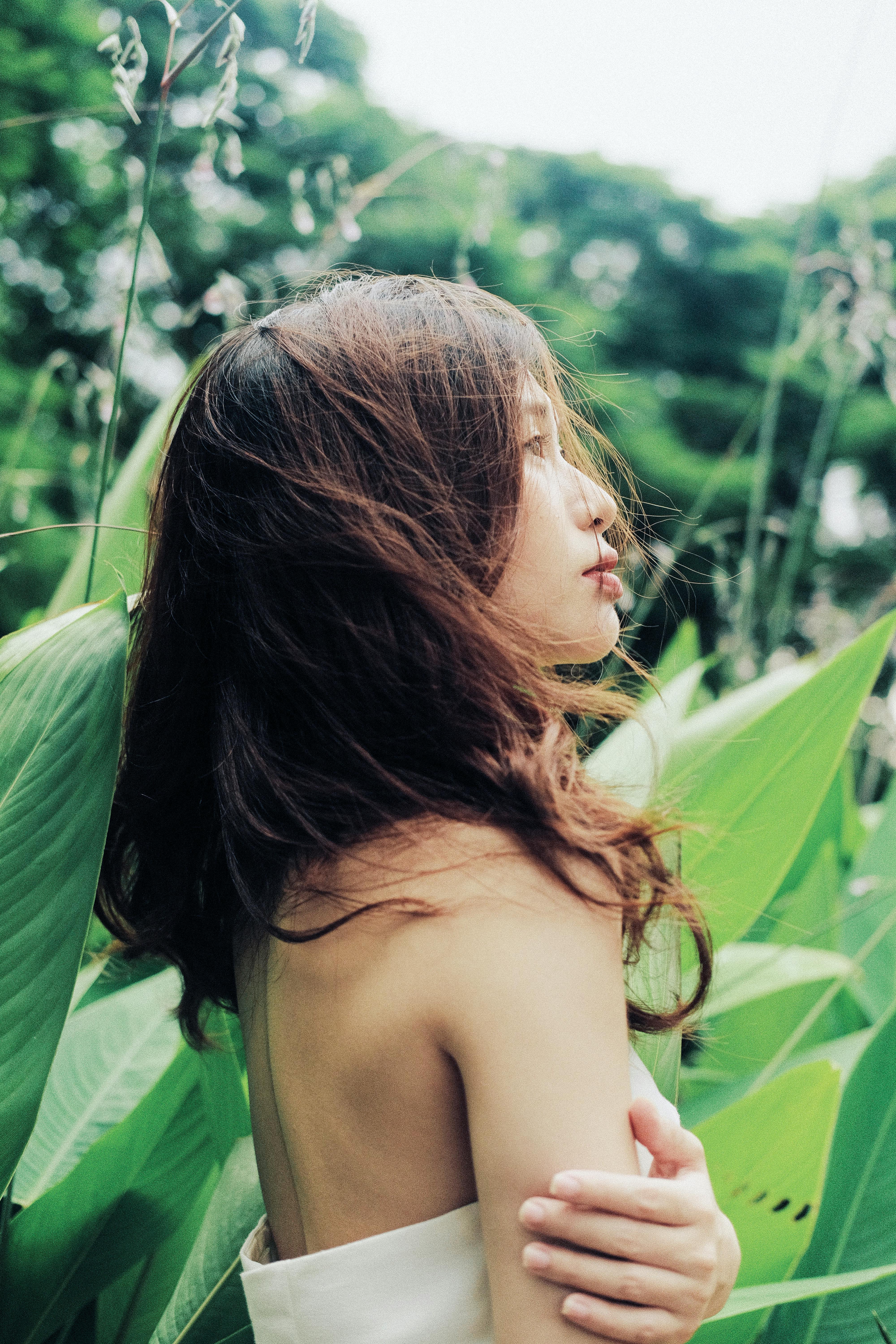 A serene young woman with brunette hair stands gracefully among lush green leaves in an outdoor setting.