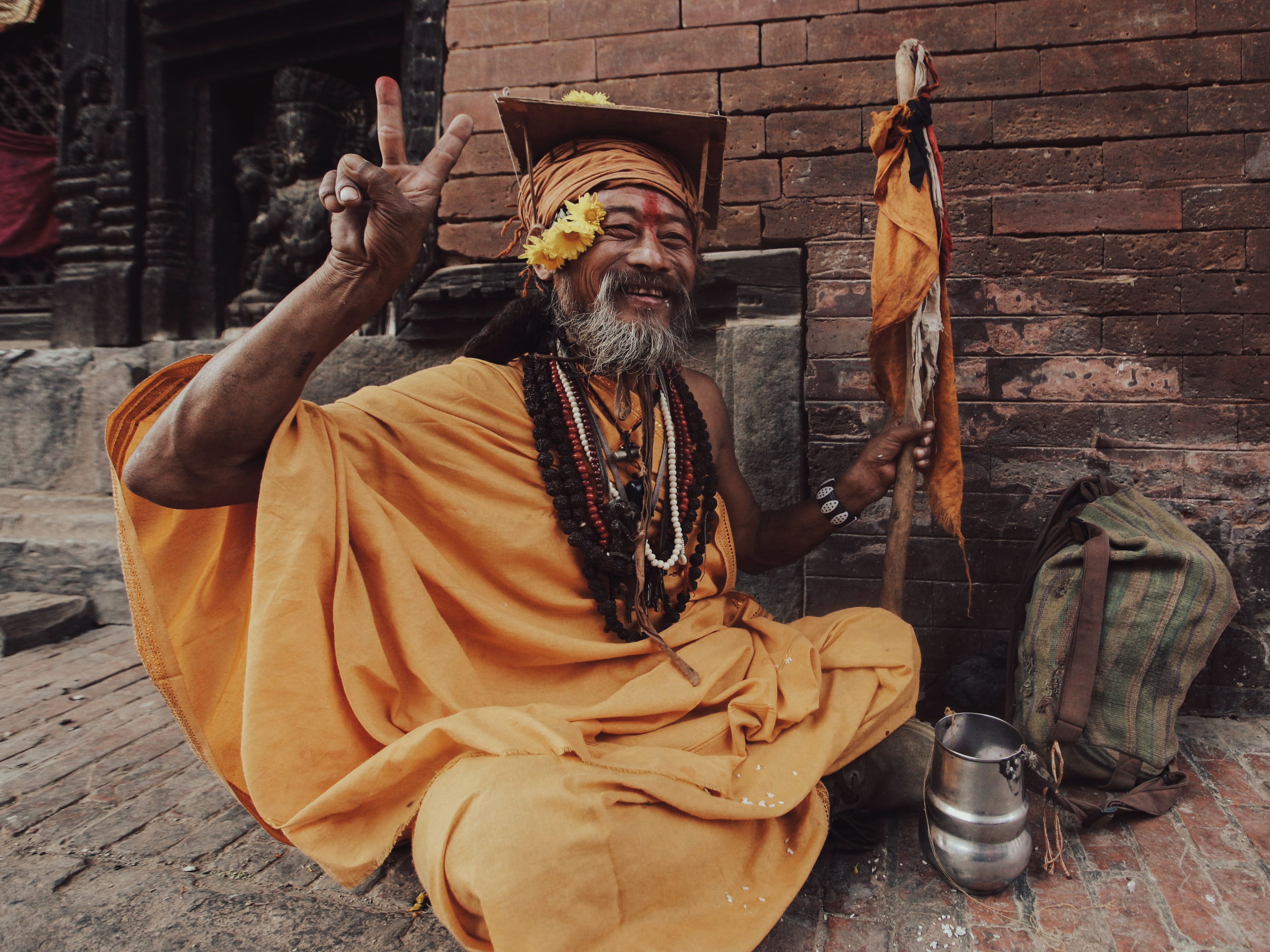 Elderly sadhu in vibrant yellow attire, sitting and gesturing peace in a spiritual setting.