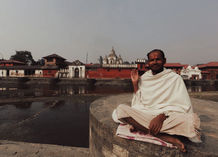 A man in traditional white robes sits near Pashupatinath Temple, Kathmandu, raising his hand in greeting.