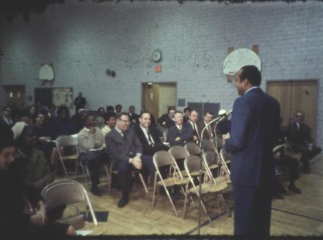 A vintage community meeting in a gymnasium with diverse audience and a speaker.