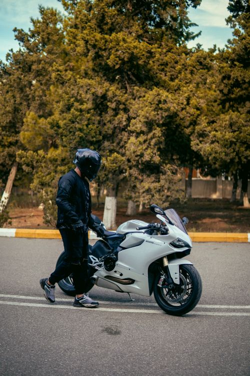 Free Motorcyclist in helmet walks beside a white motorcycle on a tree-lined road. Stock Photo